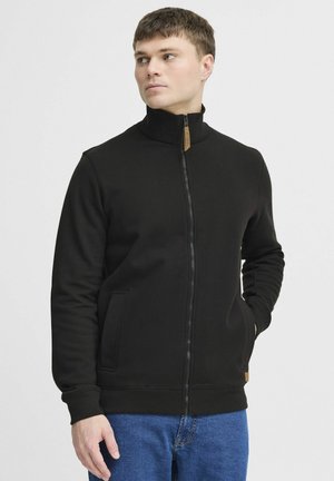 Black zip-up sweatshirt with a high collar, featuring two side pockets and a leather tag accent. Smooth texture, fitted design.