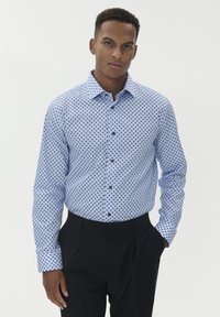 Fiatal férfi világoskék mintás ingben és fekete nadrágban áll, egyik kezével a zsebében, sima háttér előtt.