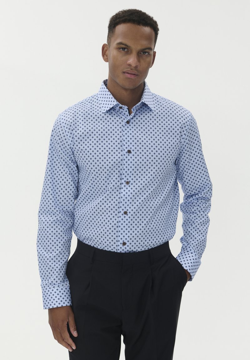 Fiatal férfi világoskék mintás ingben és fekete nadrágban áll, egyik kezével a zsebében, sima háttér előtt.