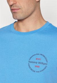 Błękitna koszulka bawełniana z okrągłym nadrukiem "NYC Tommy Hilfiger 1985" w czerwonym i ciemnoszarym kolorze, gładka tekstura, okrągły dekolt.