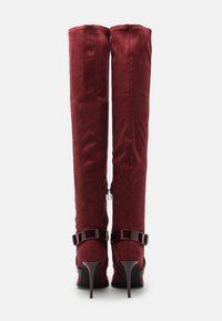 Knähöga, vinröda stövlar i mjukt tyg med glansiga lackläderdetaljer, med stilettklack och dragkedja på sidan.
