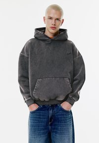 Hanorac gri supradimensionat cu buzunar tip cangur, material texturat și manșete striate. Combinat cu blugi din denim albastru. Design simplist, fără modele.