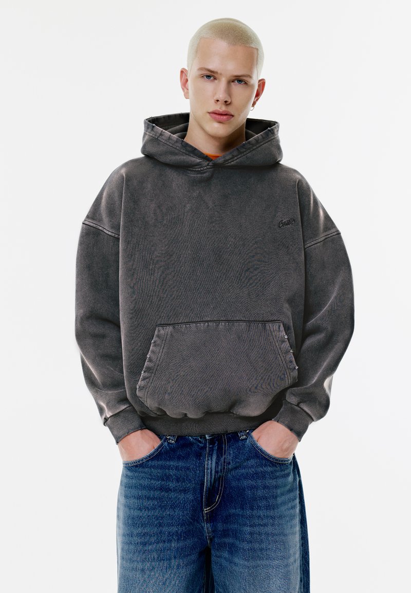Hanorac gri supradimensionat cu buzunar tip cangur, material texturat și manșete striate. Combinat cu blugi din denim albastru. Design simplist, fără modele.
