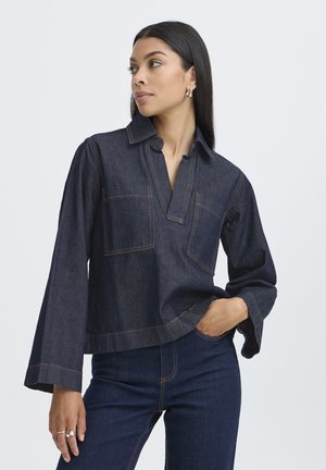 Femme aux longs cheveux foncés portant une chemise en denim bleu foncé et un jean, regardant sur le côté avec une main dans la poche, sur un fond uni.