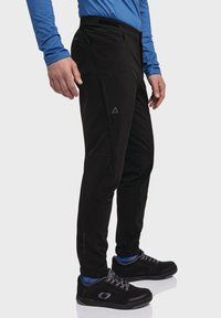 Schwarze dehnbare Hose mit slim fit, seitlichen Taschen und Logogestaltung. Trägt schwarze Schuhe, blaue Akzente sind an den Bündchen sichtbar.