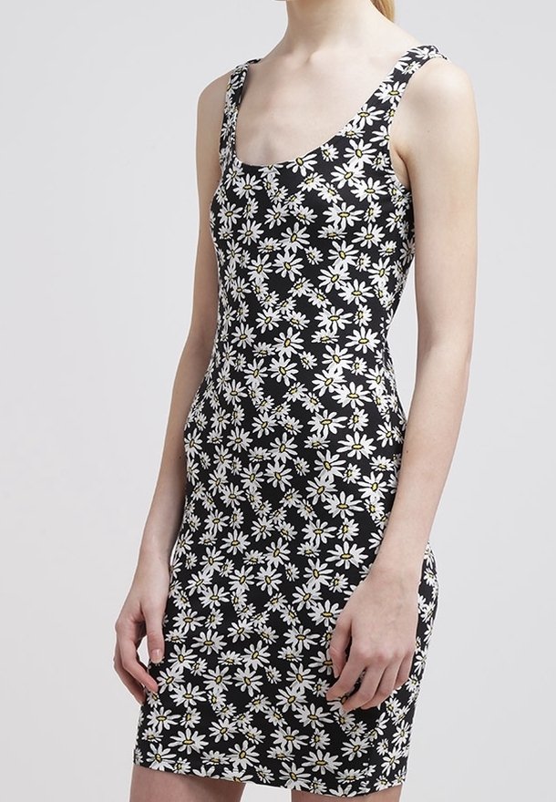 Robe noire sans manches avec imprimé marguerites blanches, silhouette ajustée, portée par une personne debout avec les bras détendus le long du corps.