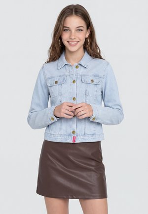 Junge Frau mit langen braunen Haaren lächelt, trägt eine hellblaue gestreifte Jeansjacke mit goldenen Knöpfen und einen dunkelbraunen Lederrock.