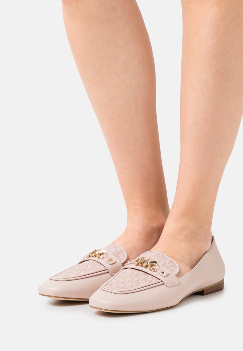 Pieds d’une femme portant des mocassins rose pâle avec des panneaux en tissu texturé et des accents décoratifs en chaîne dorée, debout sur une surface blanche.