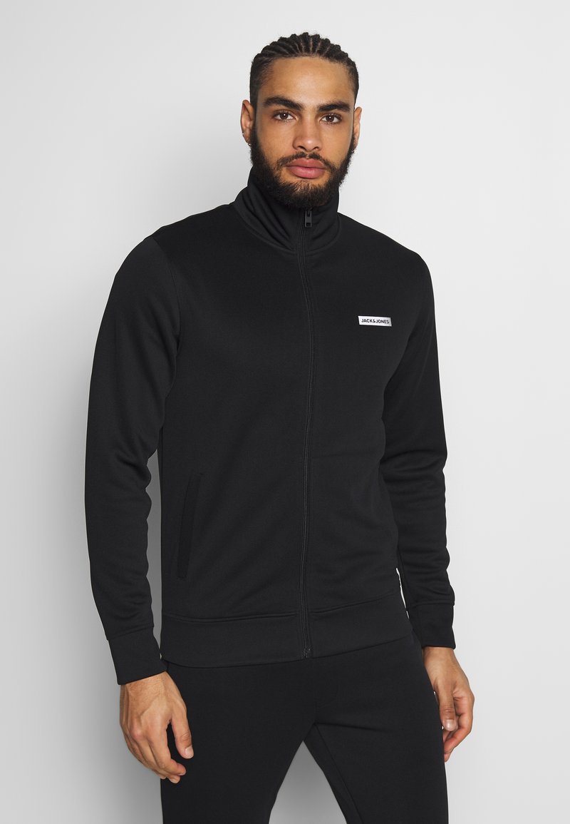 Črna zip-up bunda z visokim ovratnikom, ki ima majhno belo logotipsko nalepko in stranske žepe. Gladko blago in prilegajoč dizajn.