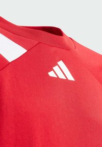 Rød idretts skjorte med V-hals, med hvite striper på skulderen og en hvit logo på brystet. Glatt stofftekstur.