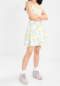 Robe fleurie avec une base blanche, ornée de fleurs jaunes et bleues. La jupe s'évase, et elle est associée à des baskets gris clair et des chaussettes blanches.