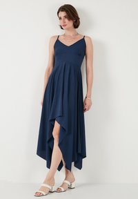 Lång navyblå klänning med V-ringning, åtsittande bodice och utsvängd kjol med asymmetrisk kant. Bärs med vita sandaletter.