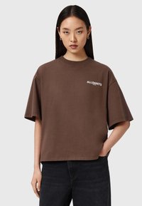 Camiseta oversized marrón con cuello redondo y mangas cortas. Presenta un logo blanco de "ALLSAINTS" en el lado izquierdo del pecho. Material de algodón.