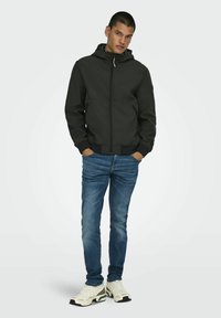 Czarna kapturowa kurtka z przednim zamkiem, połączona z jasnoniebieskimi dżinsami i beżowymi sneakersami. Kurtka ma prosty design z bocznymi kieszeniami.