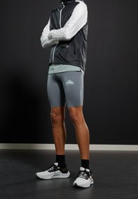 Athlète portant une veste coupe-vent noir et blanc, un short ajusté gris clair avec logo, et des chaussettes noires. Ses chaussures sont des baskets de course blanches et noires avec un accent orange.