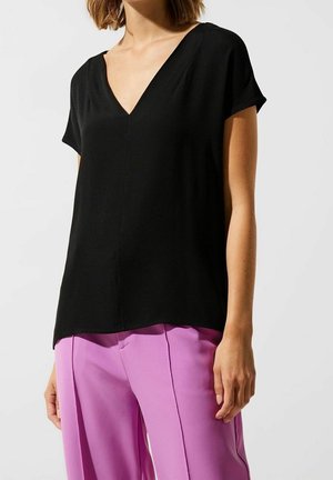 Femme portant un haut noir à manches courtes avec un col en V, associé à un pantalon violet taille haute, debout devant un fond clair uni.