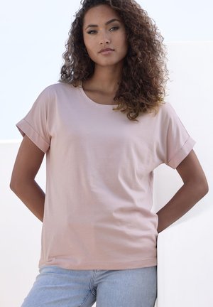 Light pink short-sleeve t-shirt in soft fabric with a round neckline, featuring rolled cuffs and a relaxed fit. Paired with light blue jeans.