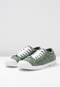 Sneakers in tela verde con punte e lacci in gomma bianca. Dettagli di cucitura notevoli e un piccolo logo rosso e verde sul lato.