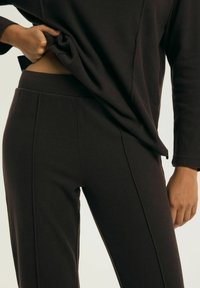 Ciemnobrązowa odzież wykonana z teksturowanej tkaniny. Bluzka jest luźna i wpuszczona w spodnie, które są dopasowane z bocznymi szwami.