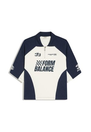 Homme portant un pantalon blanc oversize, un t-shirt noir et blanc "FORM BALANCE" et des lunettes de soleil, debout dans un skatepark au coucher du soleil avec des palmiers en arrière-plan.