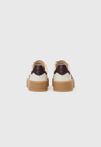Lærersneakers med en kremfarget overdel og burgunder detaljer, med en tan gummisåle og en glatt tekstur. Bakvinkel som viser logodetaljer.