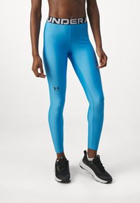 Niebieskie legginsy sportowe wykonane z elastycznego materiału, z czarnym pasem w talii, na którym znajduje się napis "UNDER ARMOUR" oraz małe logo na dolnej części nogawki.