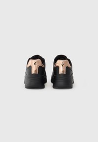 Un paio di sneakers nere con dettagli in metallo rosa oro sul tallone, viste da dietro su uno sfondo chiaro e semplice.