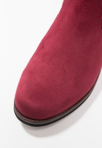 Bottine en daim rouge avec un bout arrondi, une texture lisse et une semelle marron foncé. La surface présente une finition mate uniforme.