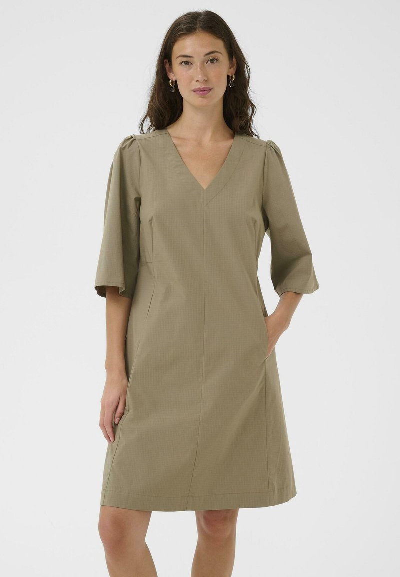 Femme aux longs cheveux bruns portant une robe olive longueur genou, manches mi-longues et col en V, debout avec une main dans la poche.