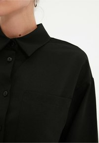 Zbliżenie na osobę w czarnej koszuli z guzikami, z kołnierzykiem i kieszonką na piersi, z widocznym fragmentem kolczyka w kształcie kółka na jasnym tle.