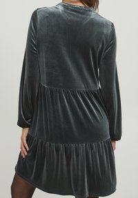 Grå sammetsklänning med långa puffärmar, en rynkad, lager-på-lager kjol och en slät textur. Har en rund halsringning och en utställd silhuett.