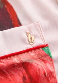 Stof met bloemenpatroon op een roze basis, met een gouden "b"-logo, rode accenten en contrasterende stiksels langs de randen.
