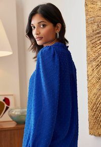 Modra teksturirana bluza s napihnjenimi rokavi in izrezom z valovito obrobo ter izbočenimi pikami po celotni površini. Model stoji na nevtralnem ozadju.