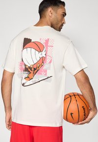 Μπεζ βαμβακερό μπλουζάκι με πολύχρωμο γραφικό που απεικονίζει ένα χέρι να κρατάει μια μπάλα μπάσκετ, με γεωμετρικά μοτίβα στο φόντο.