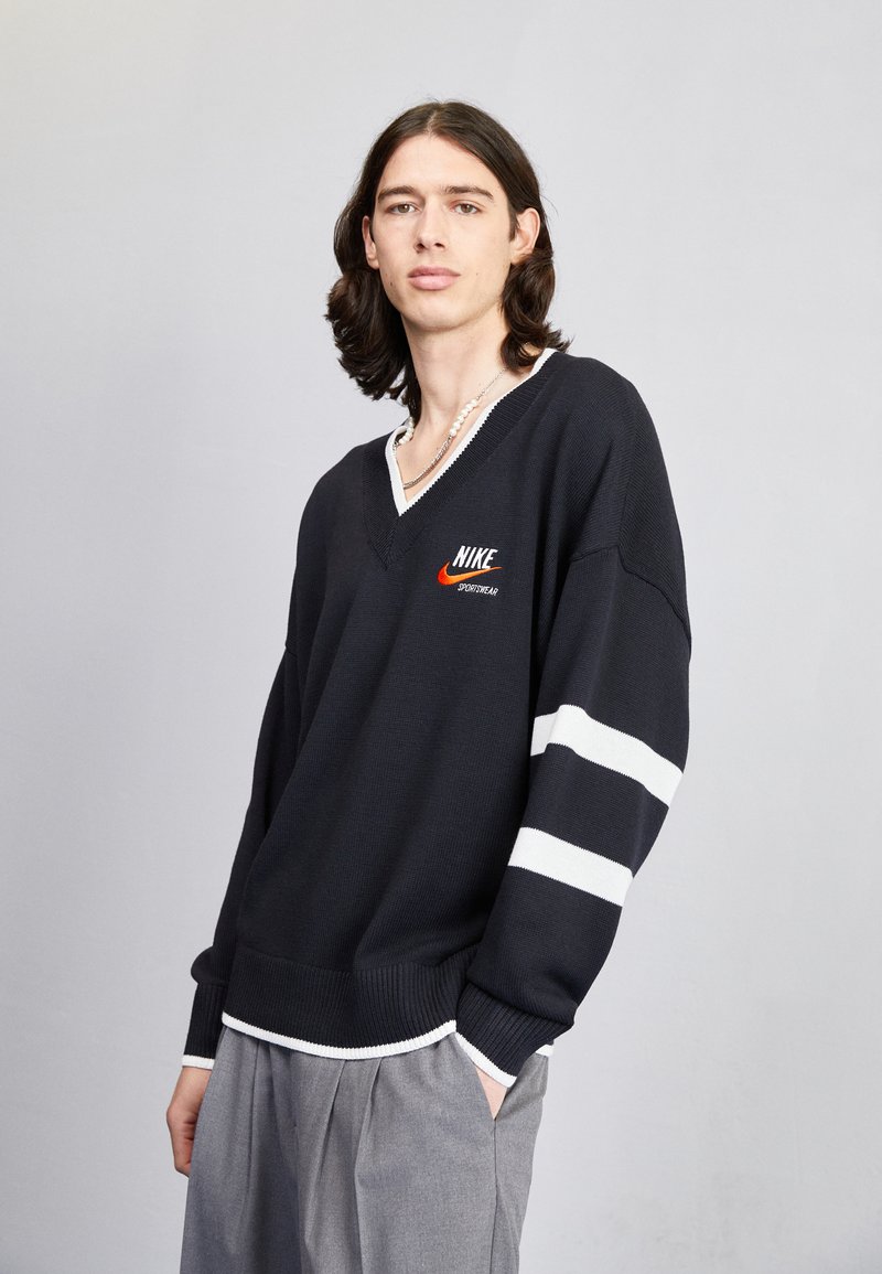 Čierny sveter s výstrihom do V a bielymi pruhmi na rukávoch a golieri. Vyšívané logo Nike v červenej farbe na hrudi. Texturovaný pletený materiál.