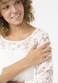 Blusa bianca in pizzo con motivi floreali, scollatura a barchetta e maniche a tre quarti, realizzata in un tessuto testurizzato e semitrasparente.