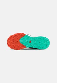 La suela de la zapatilla de trail running presenta un diseño bicolor con tacos de tracción en naranja y turquesa, un acabado texturizado y un acento central en rojo.