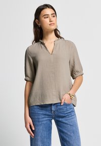 Graue, strukturierte Bluse mit V-Ausschnitt und kurzen Ärmeln, mit fransigen Kanten und lässigem Schnitt, kombiniert mit blauen Jeans.