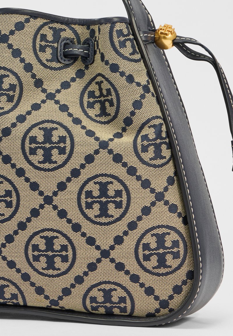 Leinenhandtasche in Beige mit navyblauem geometrischen Muster und kreisförmigem Logo. Verfügt über eine dunkle Lederverzierung und eine abgerundete Form mit oberem Verschluss.