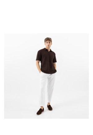 Giovane uomo che indossa una polo lavorata a maglia a maniche corte marrone scuro, pantaloni bianchi corti e mocassini marrone scuro, in piedi con le mani nelle tasche.