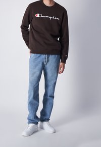 Felpa marrone con logo "Champion" bianco e rosso, abbinata a jeans in denim azzurro chiaro e sneakers bianche. Design semplice, vestibilità rilassata.