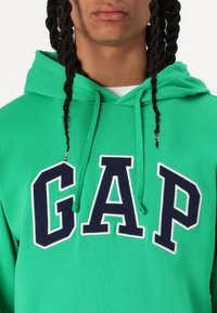 Felpa verde in cotone con un grande logo "GAP" navy contornato di bianco, con stringhe sul cappuccio e polsini a costine.