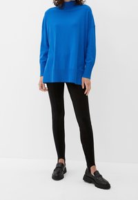 Femme portant un pull à manches longues bleu vif, un pantalon noir ajusté avec des fentes aux chevilles, et des mocassins noirs épais sur un fond blanc.