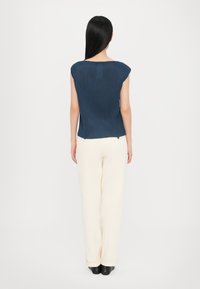Donkerblauwe ribbed top met korte mouwen, gecombineerd met crèmekleurige wijde broek en zwarte schoenen, gezien vanaf de achterkant tegen een witte achtergrond.