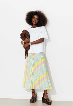 Femme aux grands cheveux bouclés portant un t-shirt blanc, une jupe plissée rayée colorée, des sandales compensées noires, tenant un sac marron à franges.