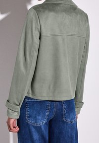 Light green suede jacket with a classic collar, hidden seams, and rolled cuffs, paired with blue denim jeans featuring back pockets.