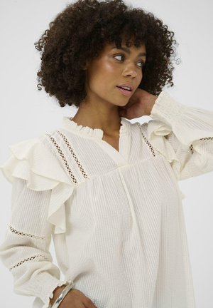Valge tekstuuriga pluus volditud varrukate, pitsdetailide ja nööbitava kaelusega. Omab voldi- ja kummist mansette mugavuse tagamiseks.