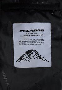 Juodos audinio etiketės su "PEGADOR OUTERWEAR" logotipu. Įtrauktos priežiūros instrukcijos ir kalnų grafika baltame fone. Tekstas pabrėžia nuotykius.