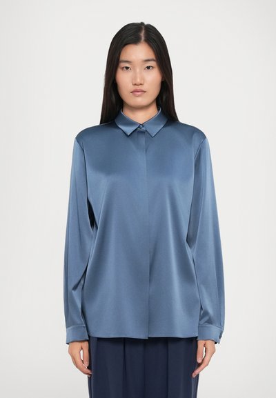 Woman with straight black hair wearing a shiny blue long-sleeve button-up shirt and dark pants, standing against a plain white background.