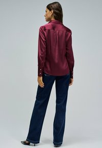 Blusa de satén burdeos con mangas largas, puños abotonados y detalle de pliegue en la parte posterior, combinada con unos vaqueros oscuros de pierna ancha.